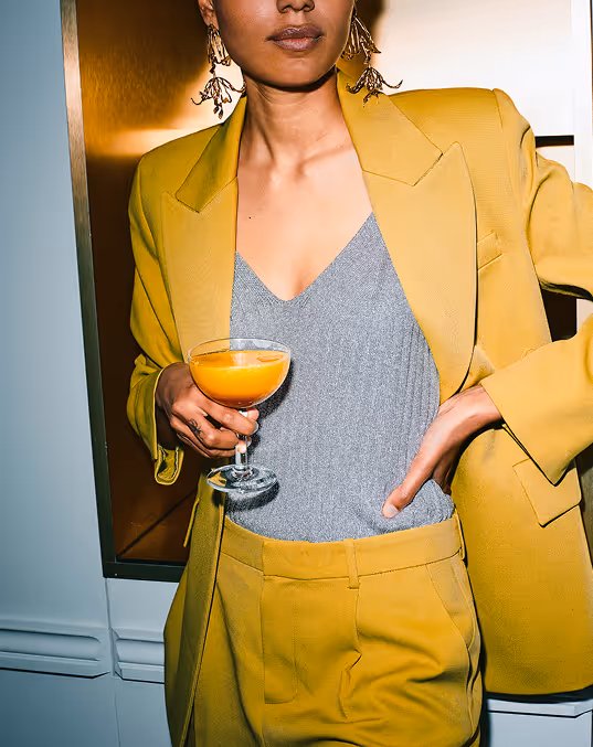 Credit: Death to Stock Person in a yellow suit holding a cocktail glass with an orange drink indoors.