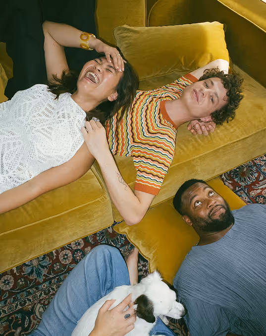 Credit Death to Stock. Three friends lying on a yellow couch and floor, one holding a white dog, all smiling and relaxed.