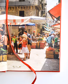 Credit Emilia Piazza. Book of image of busy outdoor market street with people shopping near fruit and vegetable stalls under awnings.