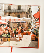 Credit: Emilia Piazza. Italian outdoor market street with people shopping under striped umbrellas and old buildings.