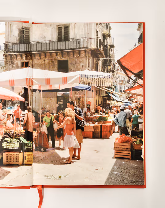 Credit: Emilia Piazza. Italian outdoor market street with people shopping under striped umbrellas and old buildings.