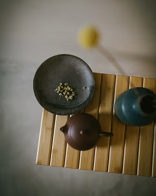 Credit Death to Stock. Overhead view of a wooden table with a dark teapot, a green vase, and a plate with dried flowers.