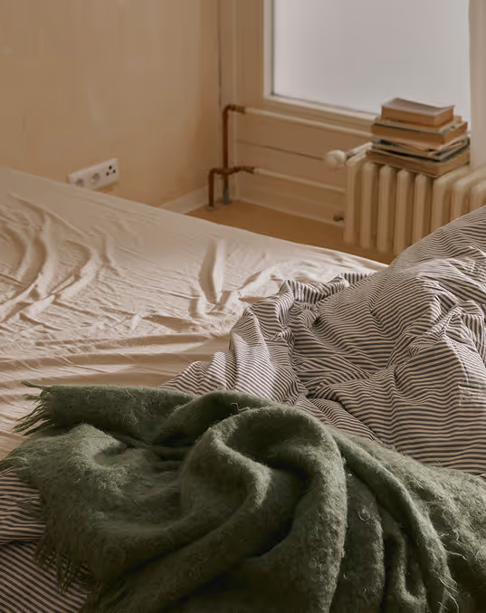 Credit: Death to Stock. 
Unmade bed with striped sheets and a green blanket, next to a window and radiator with stacked books.