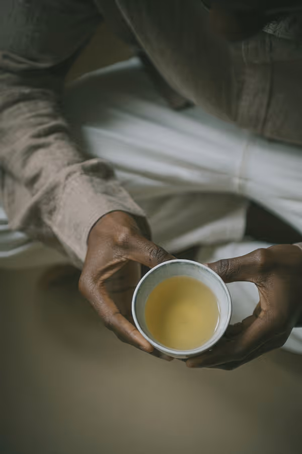 Credit: Death to Stock. Person holding a small cup with light yellow tea, wearing a beige long-sleeve shirt and white pants.